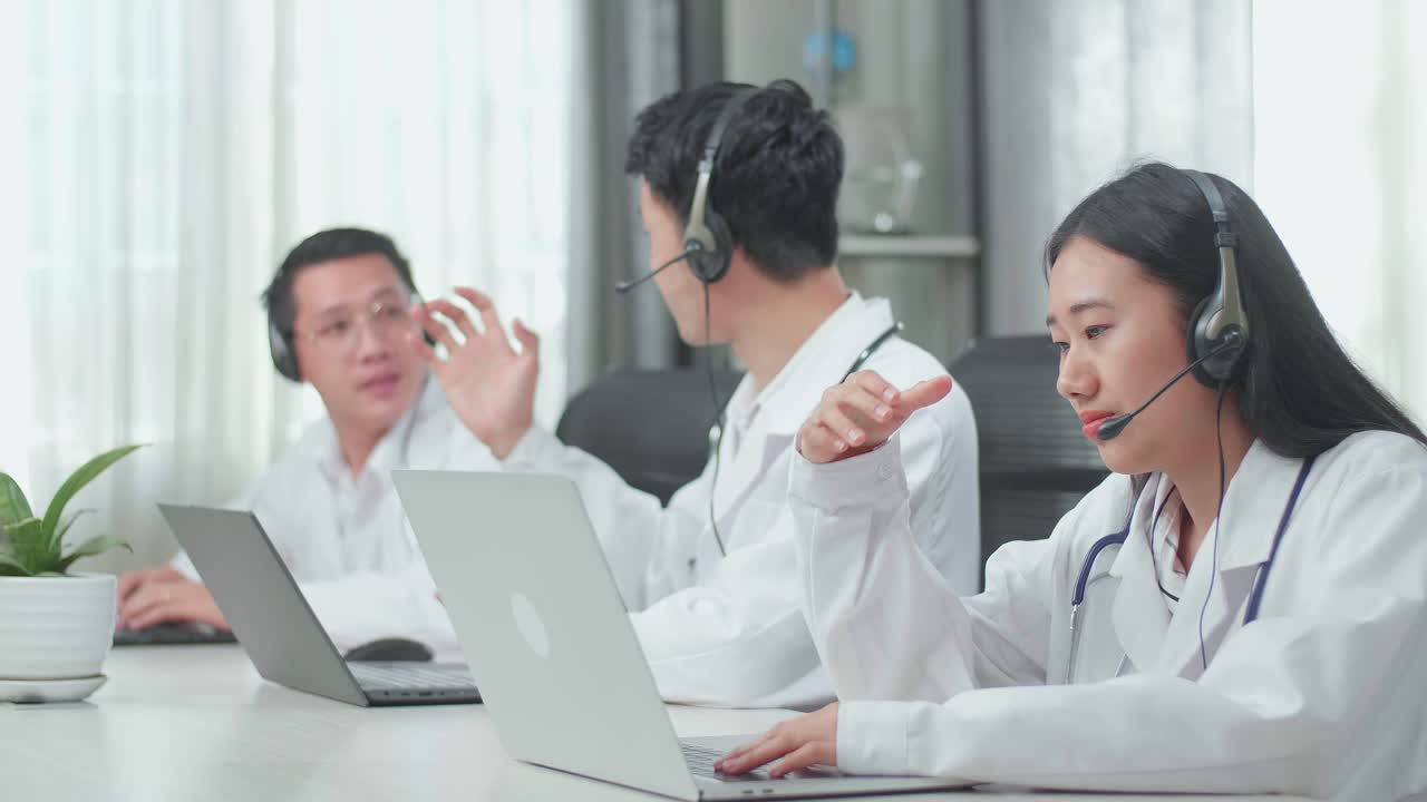 A Woman Of Three Asian Doctors With Stethoscopes Wearing Headsets Working As Call Center Agent Is Tired Because Her Colleagues Are Talking During Working At The Office