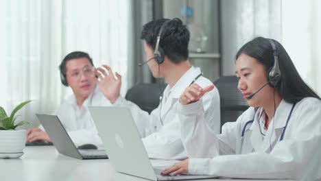 a woman of three asian doctors with stethoscopes wearing headsets working as call center agent is tired because her colleagues are talking during working at the office