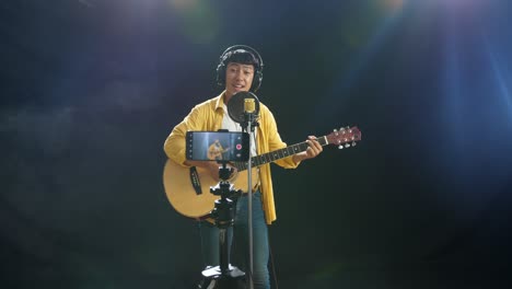 young asian boy with headphone playing a guitar and singing into a condenser microphone while recording video by smartphone on the white smoke black background