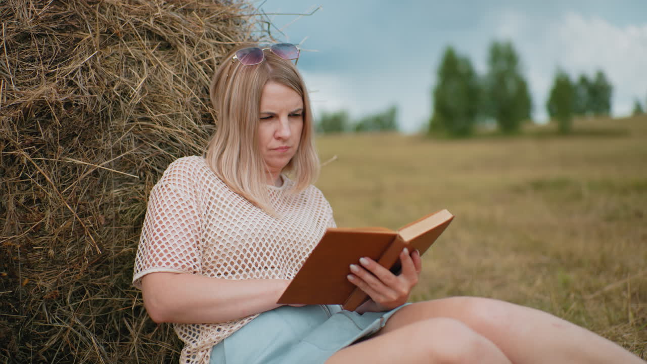 Young lady sitting on blanket, resting against hay bale, flipping book pages reflectively, legs crossed, green notebook beside her on grass, blurred background of countryside with trees and hay bales