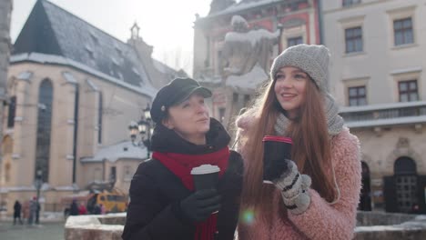 two smiling women tourists traveling together, drinking hot tea, coffee from cups on city street