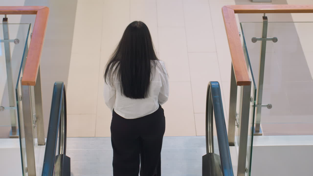 Back view of young girl with long dark hair in white shirt and black pants walking off top of escalator in well lit modern mall interior featuring clean design, glass railing, and tiled floor