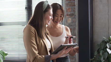 young caucasian woman and asian woman review tablet together in a business office setting