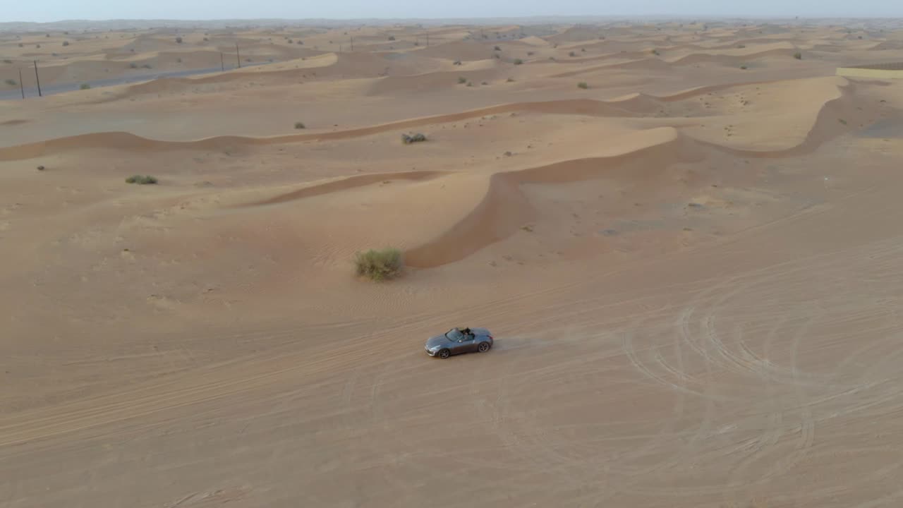 Grey convertible car rides in the desert - distance drone shot including epic dunes and sky