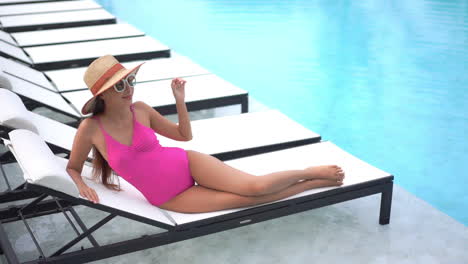 a young woman in a pink one-piece swimming suit and huge straw sun hat reclines on a poolside chaise lounger