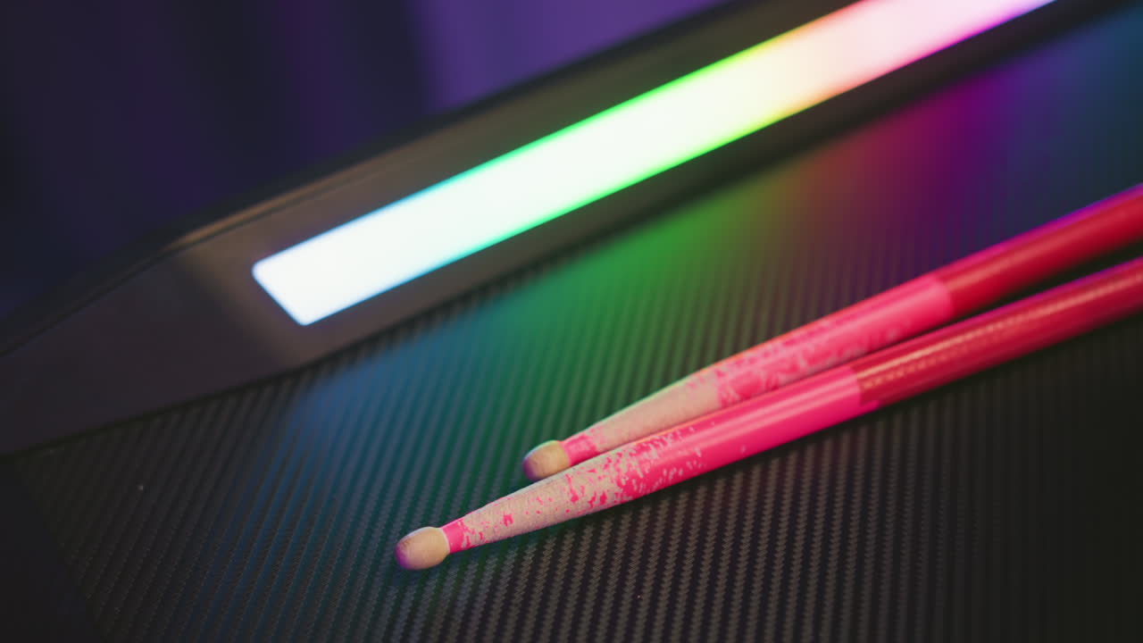 Two pink drum sticks placed on dark textured surface beside blinking multicolor LED light with soft reflections and blurred background