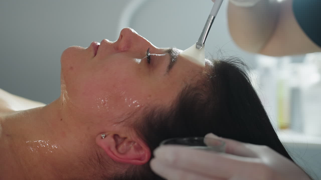 Side view of spa visitor lying eyes closed while skin expert in gloves smooths clear face gel across cheek and jawline under bright treatment light for rejuvenating facial therapy session