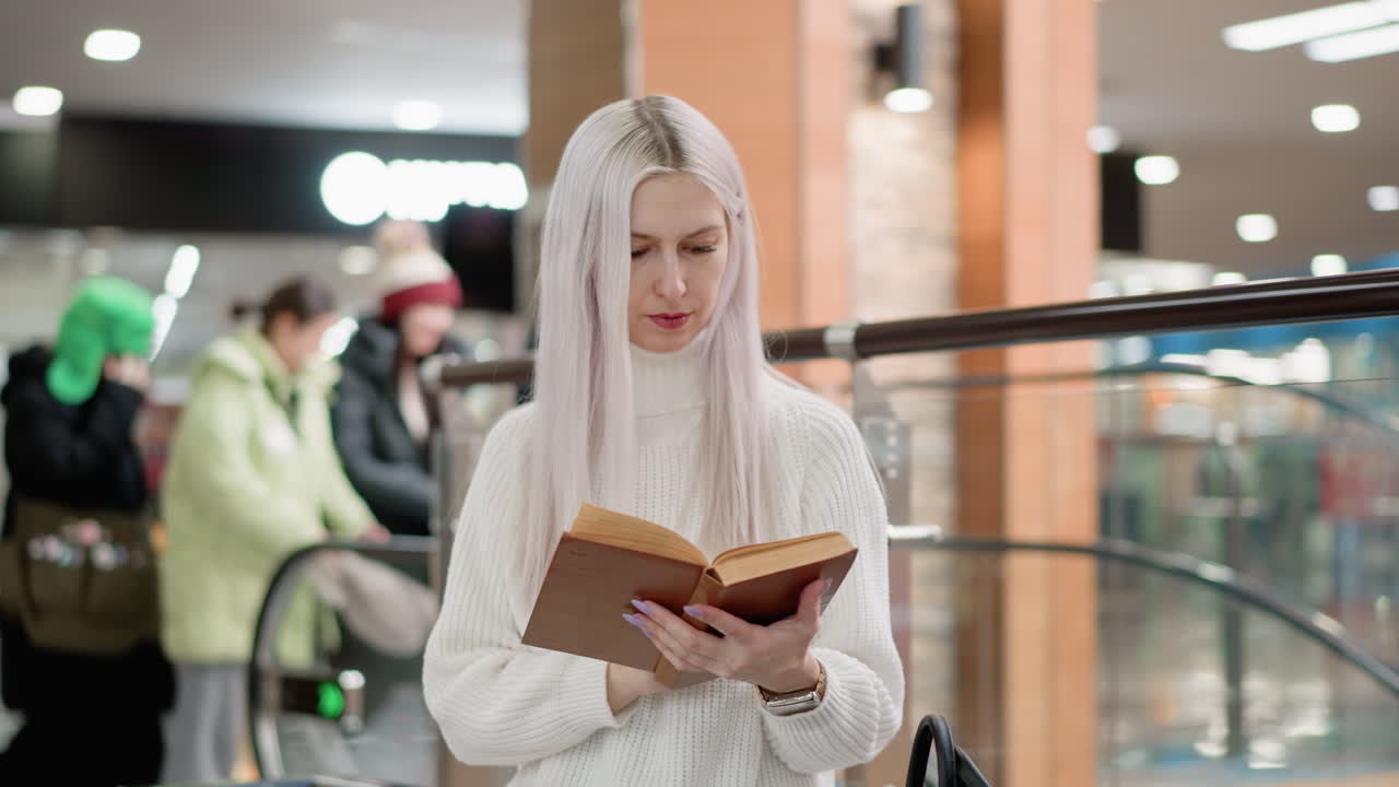 focused woman seated on bench in shopping mall reading book with intent expression flipping pages near glass railing ambient lights blurred background public space interior leisure moment
