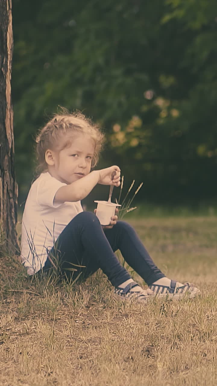young beautiful girl eats tasty yogurt and cleans mouth with hand sitting at large brown tree trunk slow motion