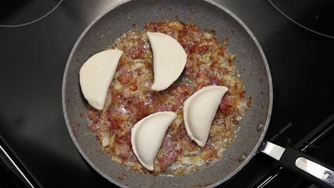 gloved hands add perogies to a pan of sizzling bacon on black stovetop