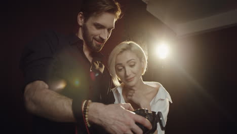 bottom view of a photographer man showing photos of photoshoot to the young blond woman model on the little camera screen in the dark studio