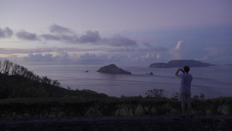 Male-using-his-phone-to-take-pictures-to-take-pictures-from-viewpoint-looking-out-towards-beautiful-ocean-and-islands