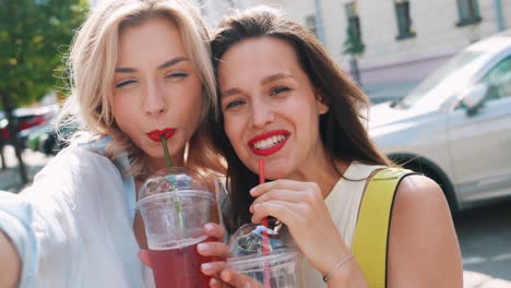 two happy friends taking a selfie while enjoying drinks on the street