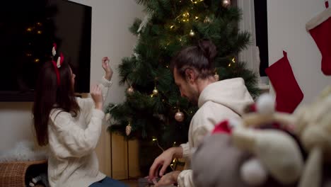 vista trasera de una chica morena feliz junto con su novio decoran el árbol de año nuevo y se preparan para la navidad en su acogedora casa en invierno