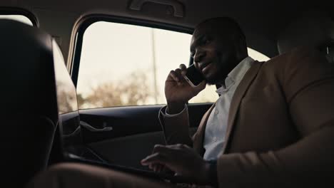 happy and confident man businessman with black skin color in a brown jacket communicates on a smartphone and types on his laptop while driving in the passenger seat in a modern car outside the city