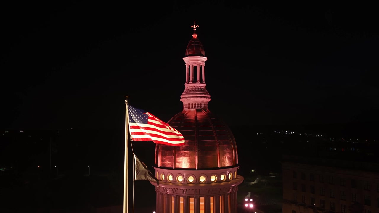 Bandiera americana sventola accanto alla cupola di rame illuminata della New Jersey State House a Trenton, New Jersey, di notte. Lanterna luminosa e colonne si ergono contro il cielo serale scuro. Veduta aerea da drone