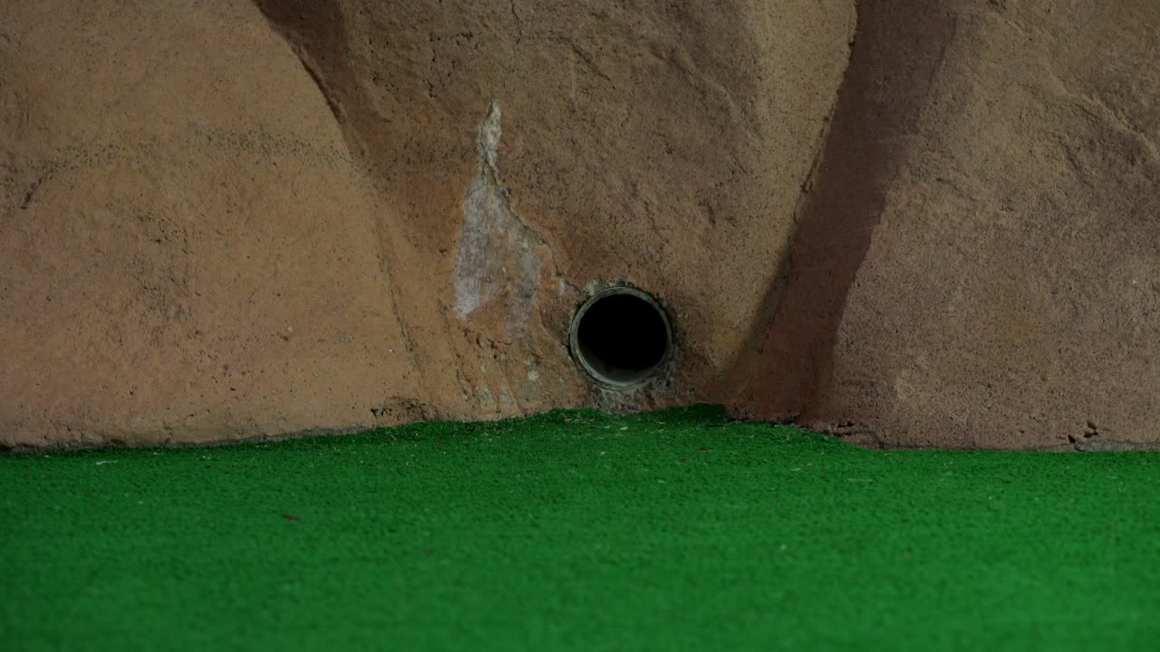 una pelota de minigolf naranja sale de una tubería en una pared y rueda y no alcanza el hoyo