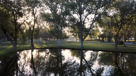 Flying-over-beautiful-park-pond-with-reflection-of-trees-toward-setting-sun