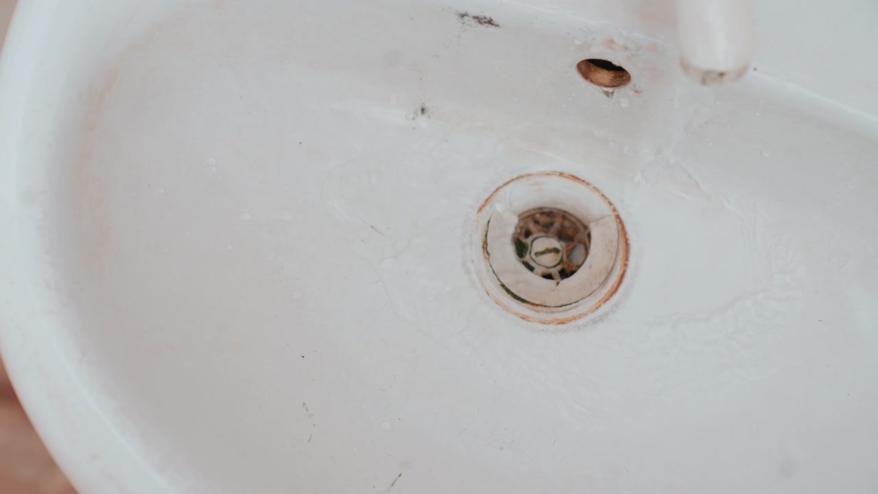 Gros plan du drain d'un évier en porcelaine taché montrant un anneau de rouille, des résidus de savon et de la saleté. Le cadrage serré capture l'émail usé, l'accumulation de minéraux autour du drain, le petit trou du robinet, la surface texturée de la porcelaine.