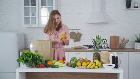 joyful woman taking out fresh fruit from paper bag in the kitchen. organic fruit and vegetableson the table and beautiful female indoors. healthy lifestyle. vertical video
