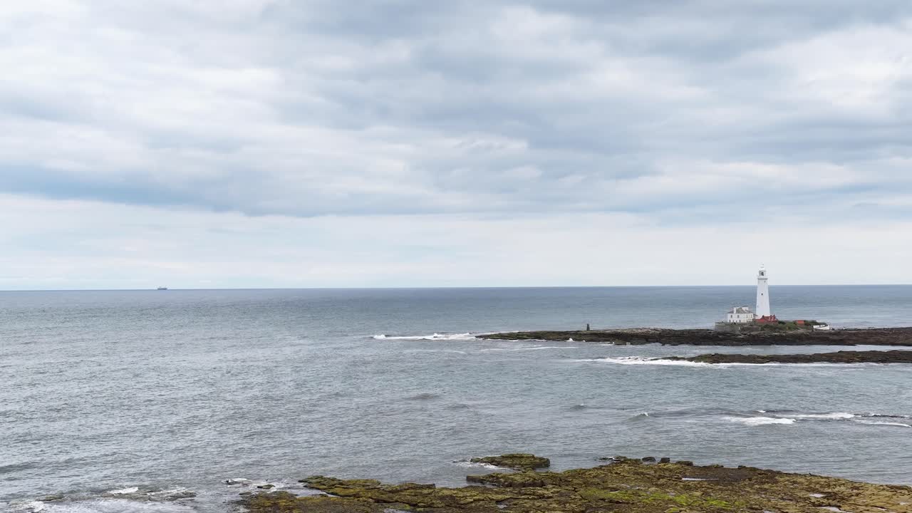 Wide shot of a white lighthouse on a rocky shoreline under cloudy skies, with smooth horizontal camera pan revealing more of the coastline and open sea