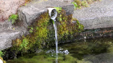 Agua-Que-Fluye-De-Un-Pico-De-Plástico-Sucio-Y-Cubierto-De-Musgo-En-Una-Fuente-De-Piedra