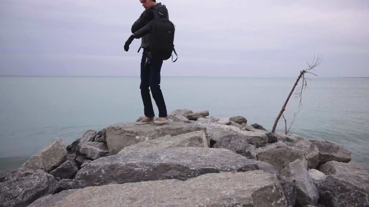 Man Sitting And Walking On The Rocky Shore In Canada While Enjoying The Sea - Wide Shot