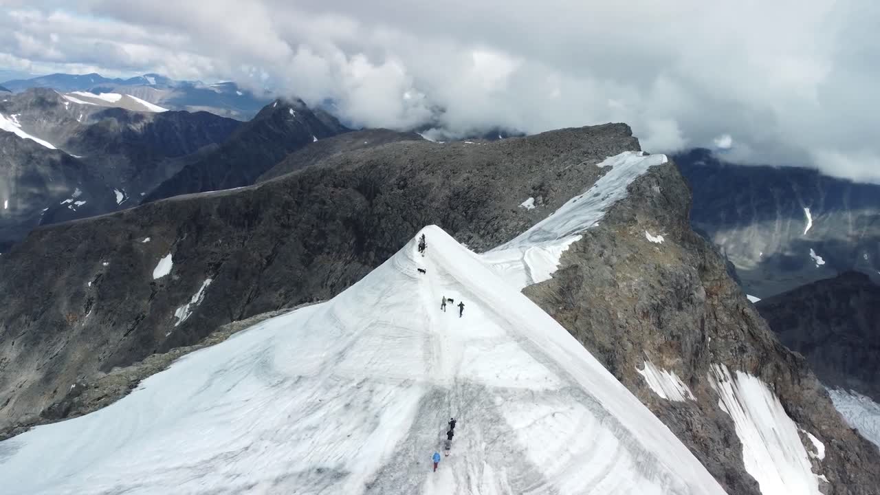 Aerial drone footage showing a group of people in winter clothing walking up a white snow covered mountain ridge or hill during winter cloudy day in sweden Kebnekaise mountains. Large mountains seen.
