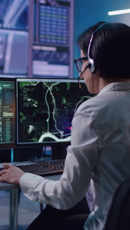 Female-employee-browsing-video-on-computer-and-talking-on-headset-while-working-in-office-of-surveillance-service.-Vertical-shot