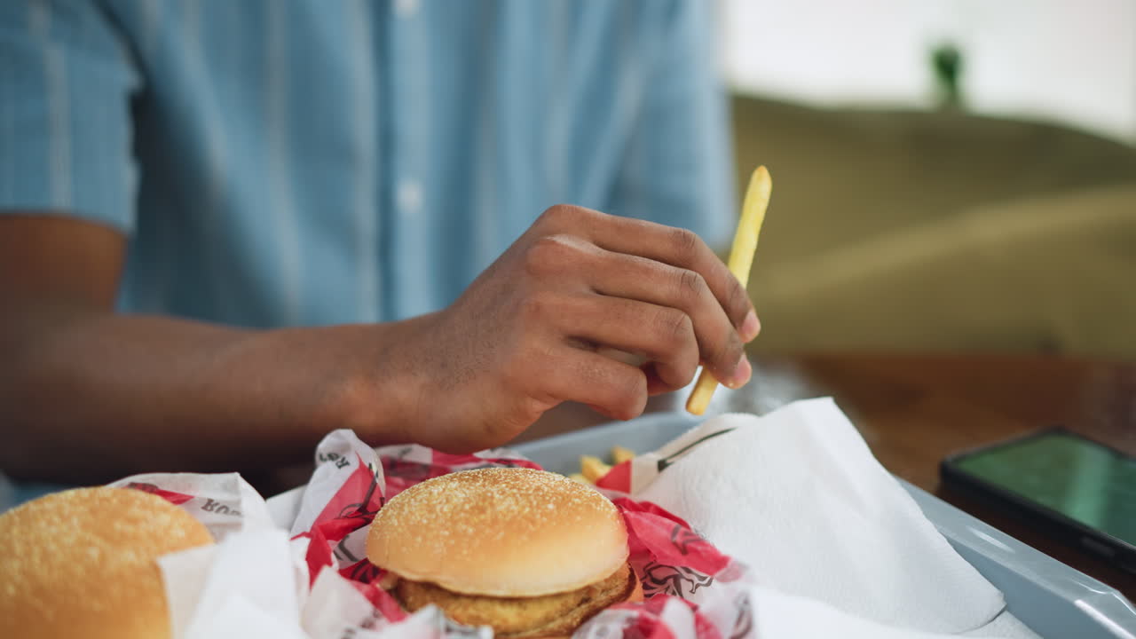 Vista ravvicinata di un hamburger su un sacchetto KFC con un bastoncino di patatine sul tavolo accanto al telefono, sfondo leggermente sfocato, attenzione ai dettagli del cibo e al dispositivo che suggerisce una pausa pranzo informale e un appetito rilassato