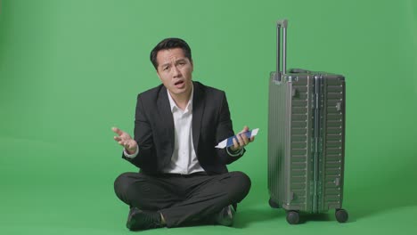 full body of wondering asian businessman traveler with luggage holding passport and sitting doubtfully in the green screen background studio