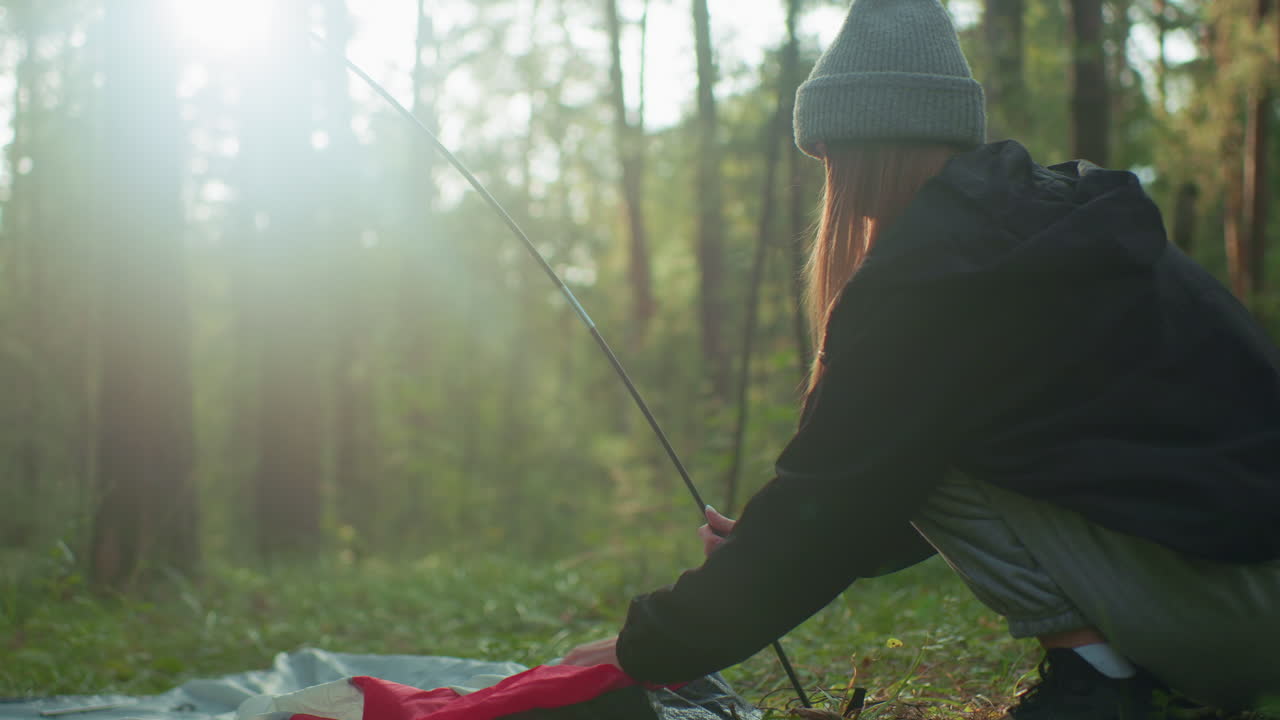 Side view of girl squatting in forest holding flexible tent pole while placing tent fabric around base, sunlight beams through tall trees casting warm glow