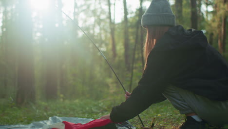 Side view of girl squatting in forest holding flexible tent pole while placing tent fabric around base, sunlight beams through tall trees casting warm glow