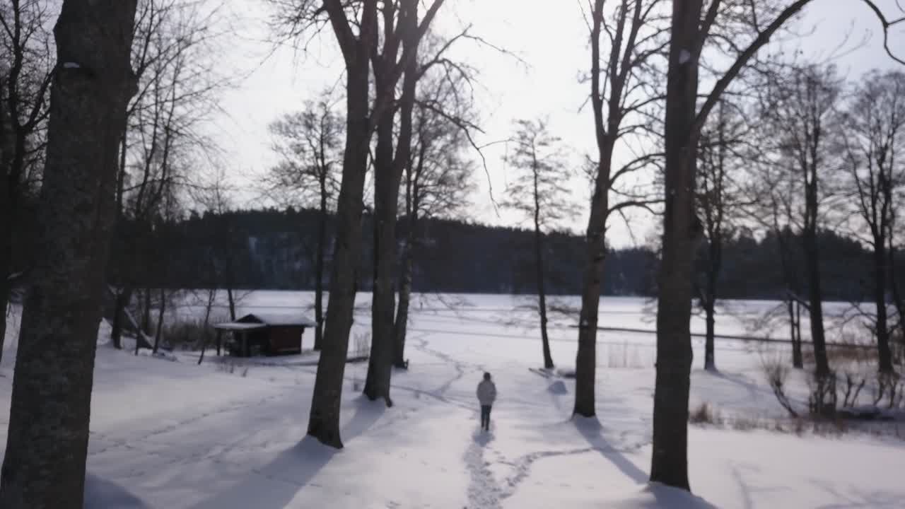imágenes aéreas que siguen a una chica rubia caminando hacia una cabaña al borde de un lago congelado en un hermoso paisaje invernal
