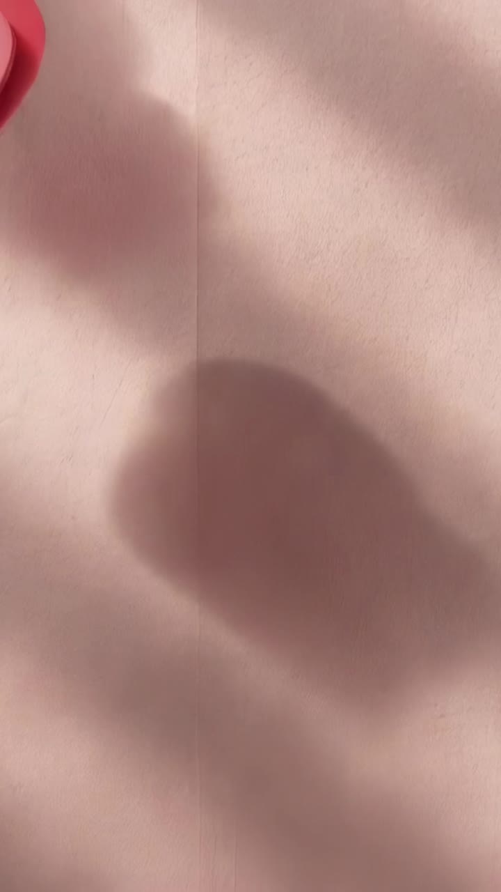 Vertical video: Shifting sunlight revealing layered red-pink heart on pale-pink sheet, copy space