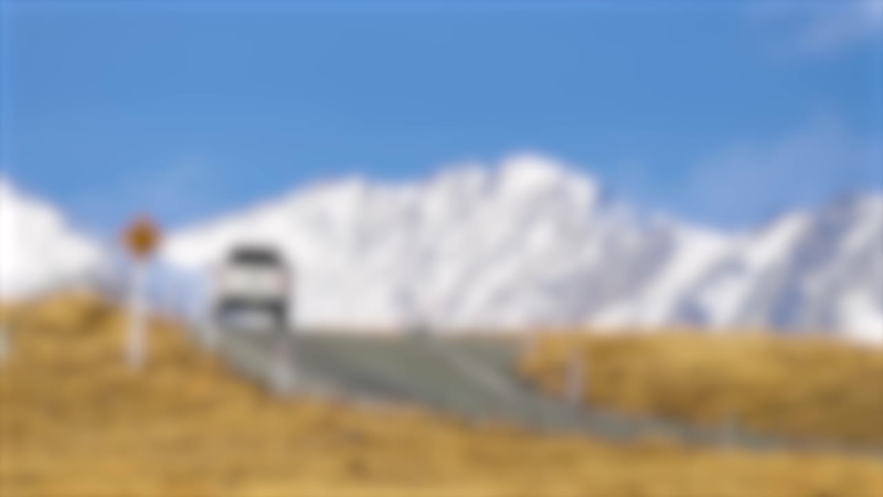 A white vehicle travels along a winding road bordered by golden grasses, with snow-covered mountains and blue sky in the background. Daylight, static wide shot