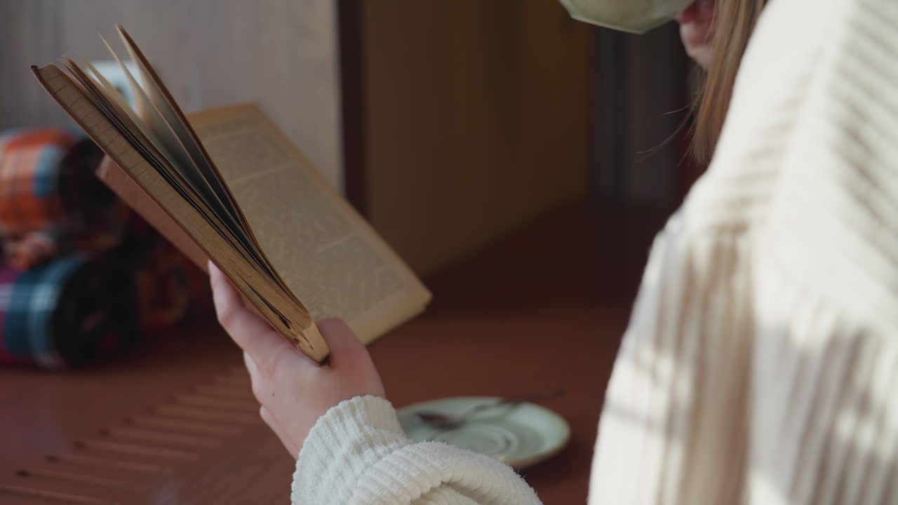 Partial view of woman wearing ribbed sweater reading vintage book while holding ceramic coffee cup in warm indoor setting with soft lighting and plaid blankets in background