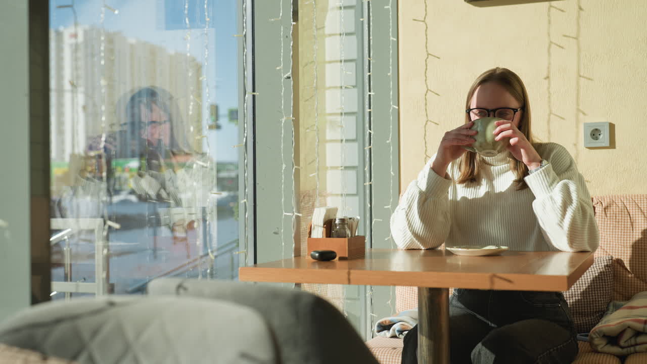 Close up of modern cafe table with woman seated drinking tea, soft sunlight casting shadows, background shows snow covered cityscape through window with gentle reflections and decorative lights