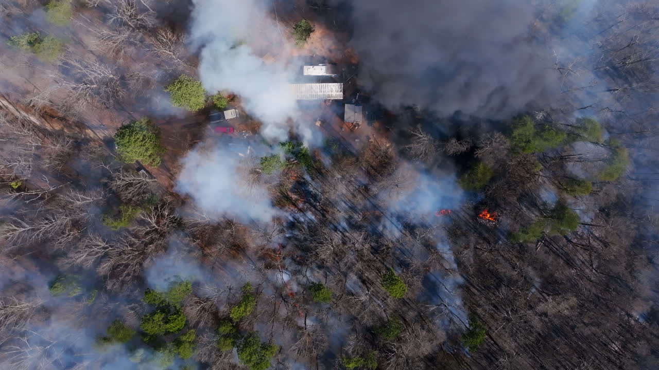 Top down aerial footage of a house in the forest with a wildfire burning all around it.