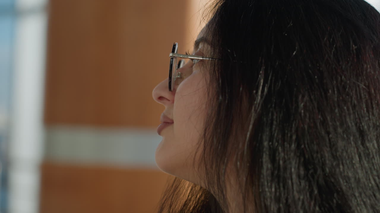 Close up of fair skinned woman with long dark hair and eyeglasses smiling gently in natural light, standing in bright modern environment with warm tones and thoughtful expression