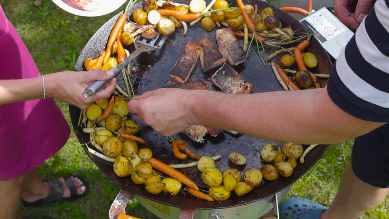 Footage shows a man and a woman serving and taking themselves salmon, potatoes and veggies from a grill or a wok grill during the summer time while the sun is shining. They life the food with spatula.
