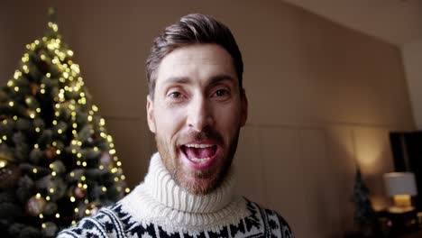 close up of happy young male in positive mood video chatting near glowing decorated xmas tree