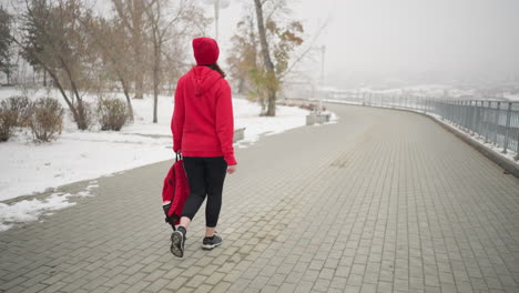 sportswoman wearing red jacket with headphones around neck walking along interlocked pathway holding a bag in her left hand near iron railing in a serene park with snowy ground and foggy atmosphere