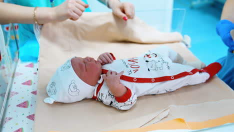 tiny little child in a cap and sleepsuit lies on the table laid with sheets. nurse in latex gloves puts on warm sock on baby's feet.