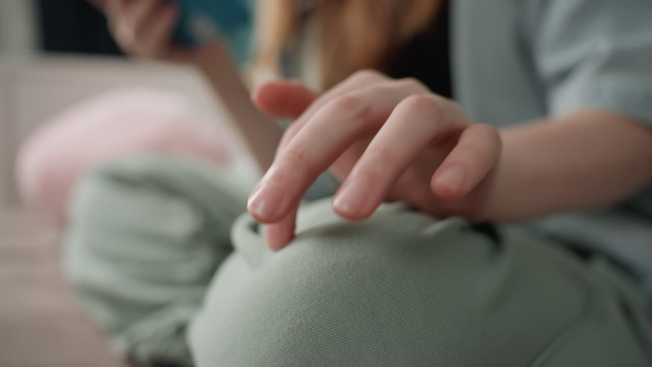 Caucasian girl mindful hand resting on knee while holding tablet, gentle fingertip tapping and slow breath, textured cushion and relaxed domestic setting, subtle motion and self soothing focus