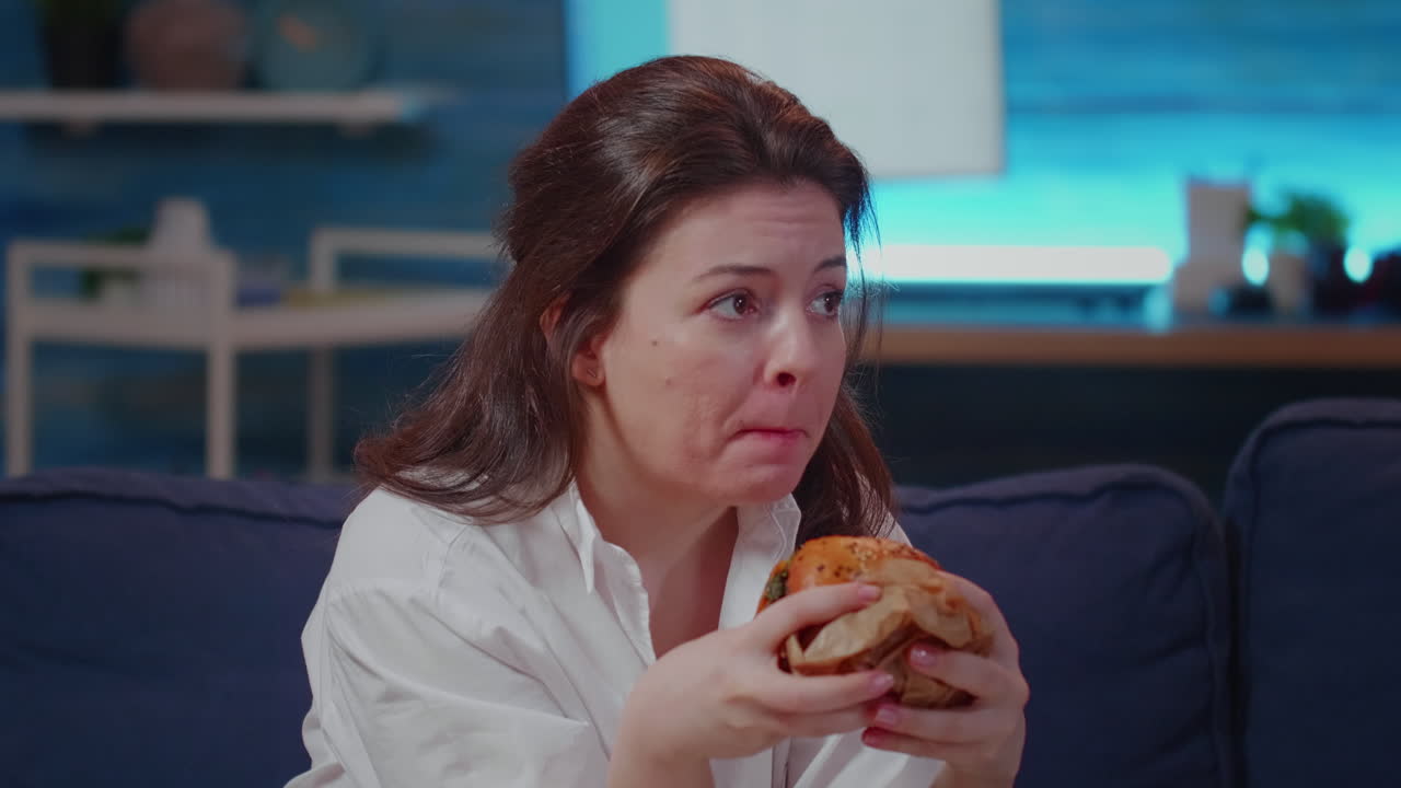 Caucasian woman taking bite from hamburger in living room