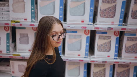 aerial close-up of young woman walking by a store shelf filled with bedding products, she observes different designs, evaluating comfort and style options, retail shopping experience