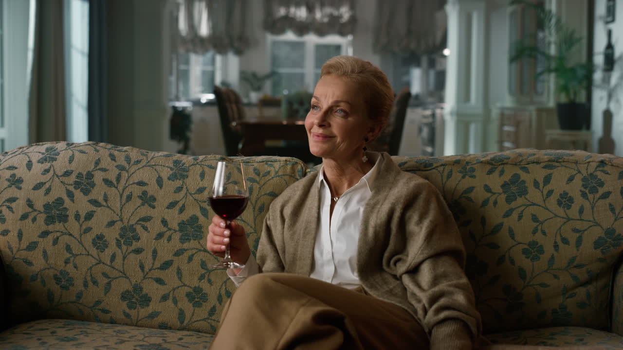 Dreamy old woman tasting red wine glass in vintage living room