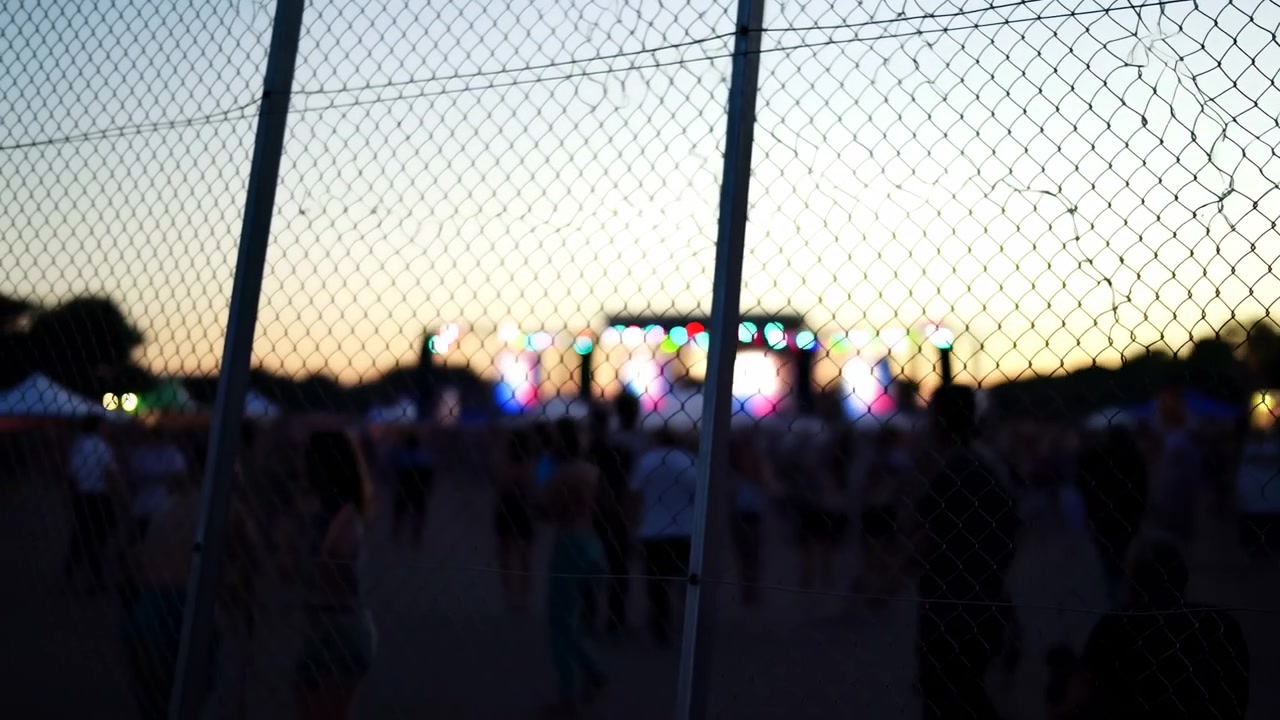 Rete metallica e pali di recinzione verticali rimangono nettamente prominenti mentre visitatori sfocati passano dietro di essi, mentre una luminosa illuminazione per eventi risplende in lontananza al crepuscolo