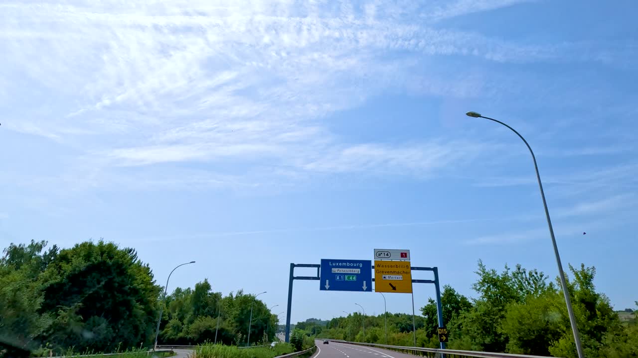 A vehicle travels along a tree-lined highway in Luxembourg, passing directional road signs under bright daylight with clear blue skies and scattered clouds